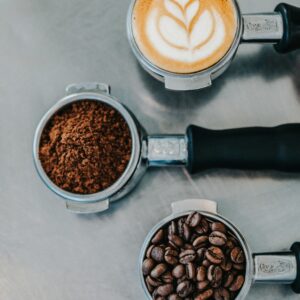 Vue de dessus de trois porte-filtres espresso contenant un latte avec mousse latte art, du café moulu et des grains de café sur une surface métallique.